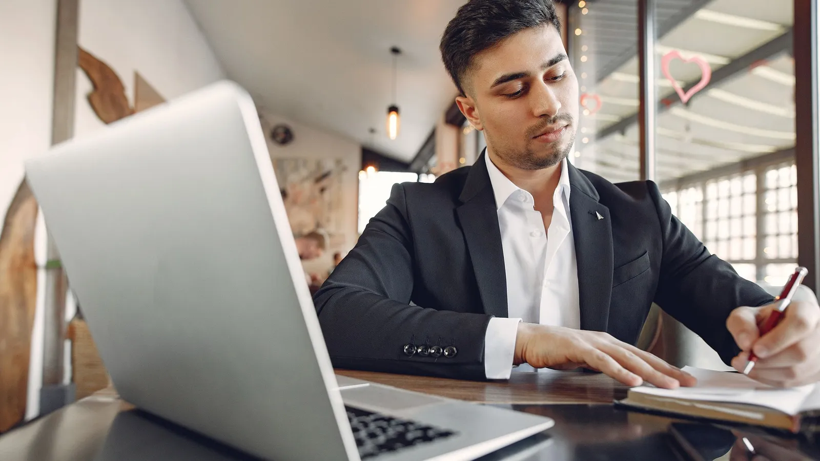 A young South Asian man in a suit works at a laptop and takes notes at an office table.