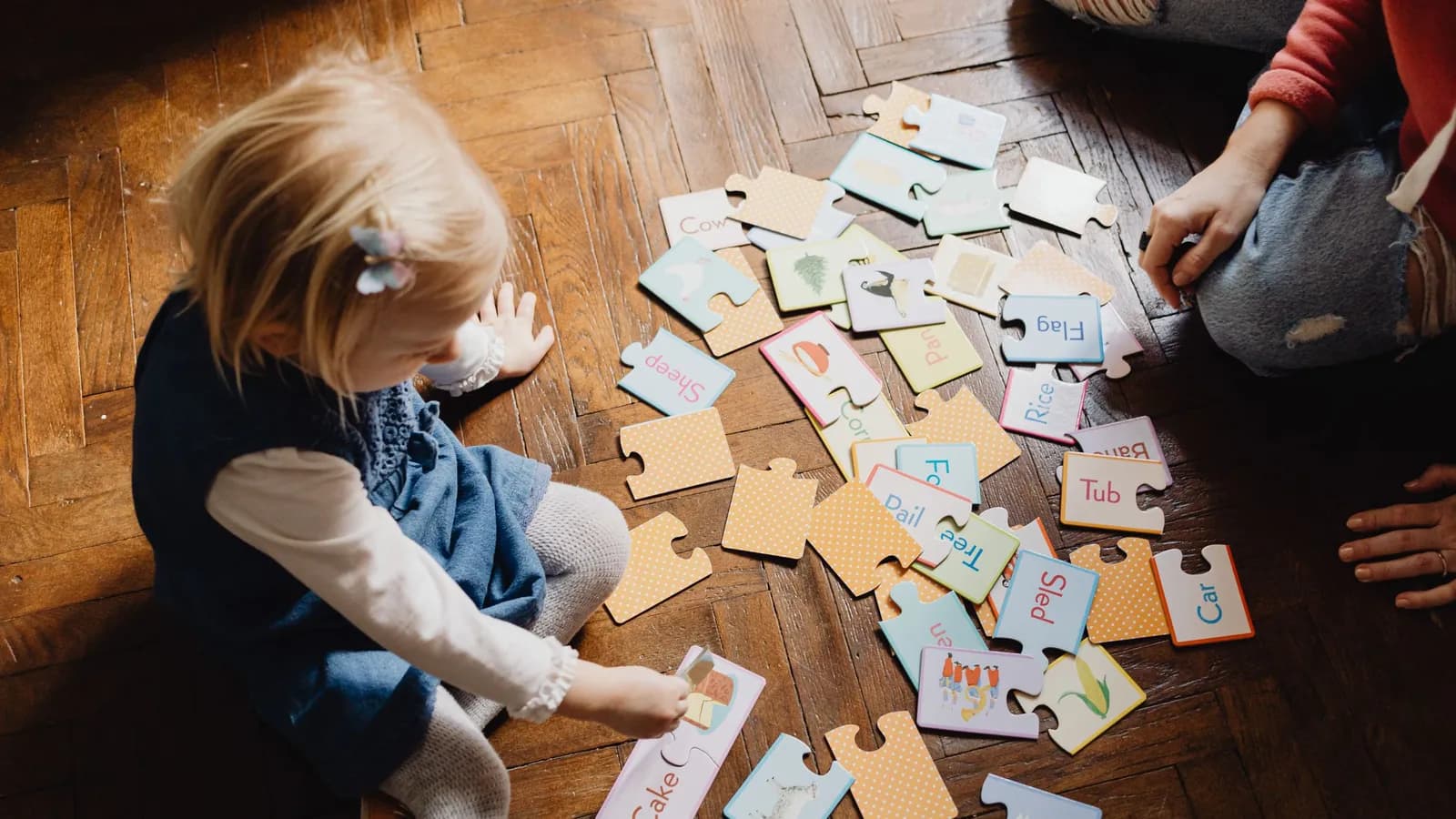 A small child plays on a wooden floor with puzzle pieces showing English words and pictures.