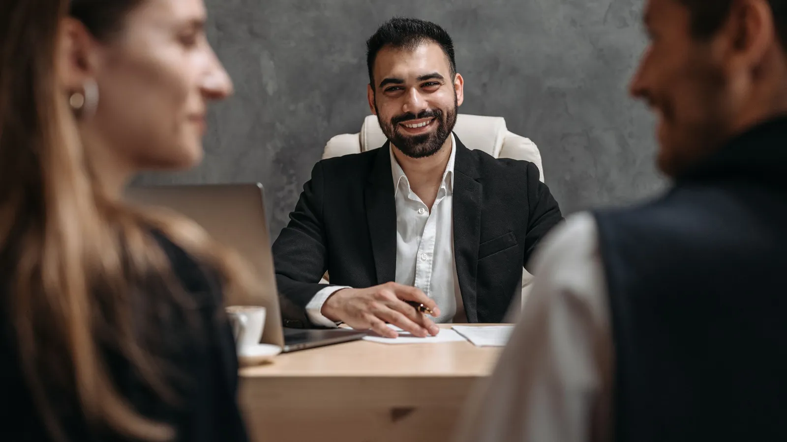 A smiling South Asian man in a suit sits across a table from two people in an office interview.