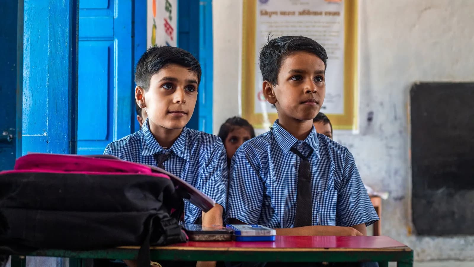 Two Indian schoolboys in uniform and ties sit at a desk listening in class.
