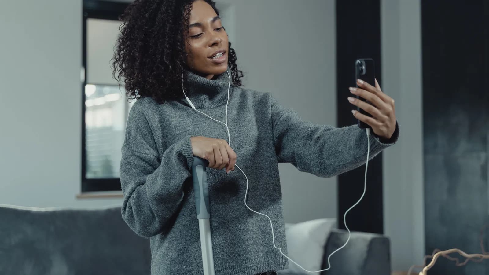 A woman with earphones talks to her phone, held up in front of her, while cleaning the living room.
