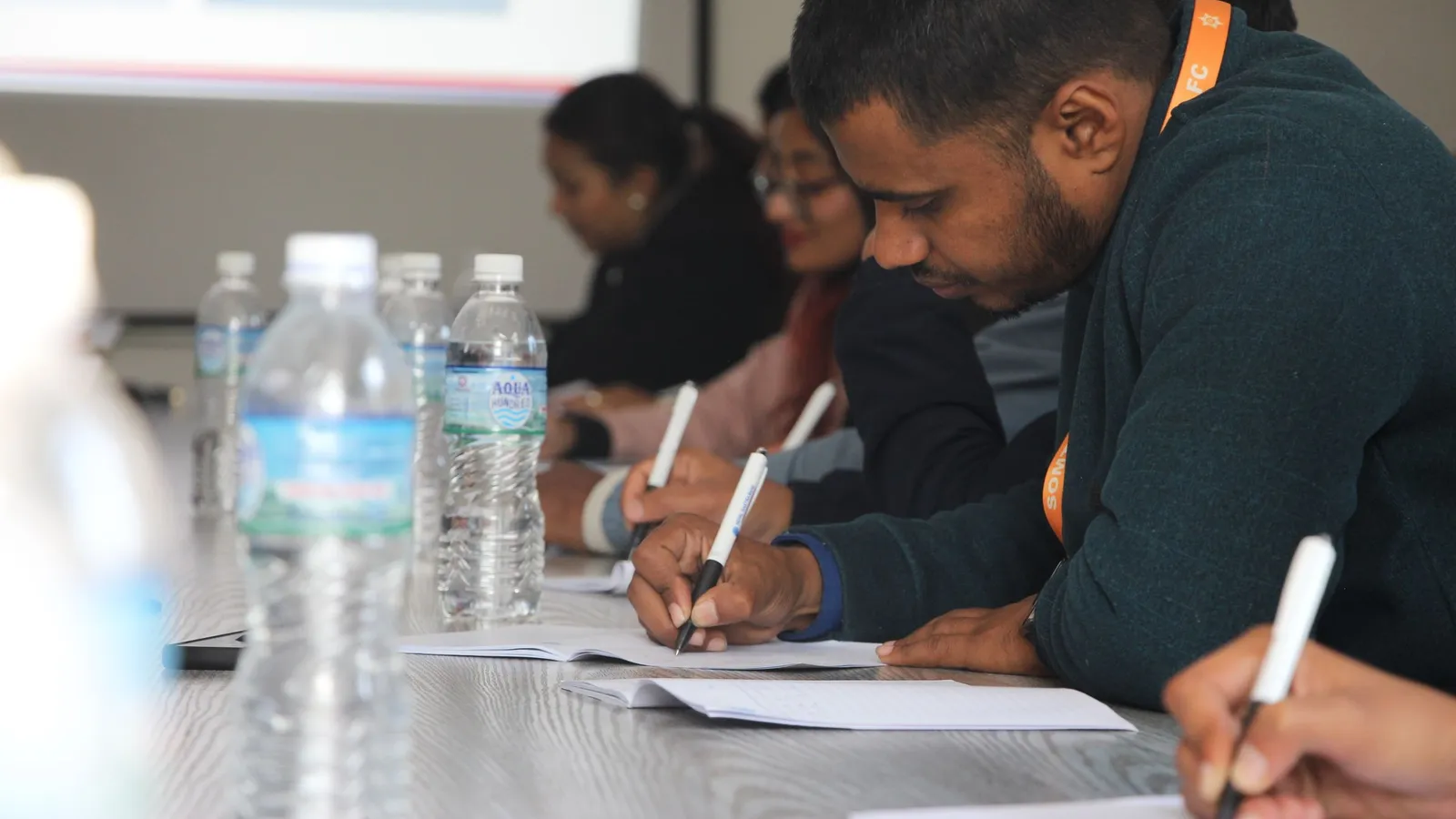 Adult trainees sit along a table, taking notes during a training session.