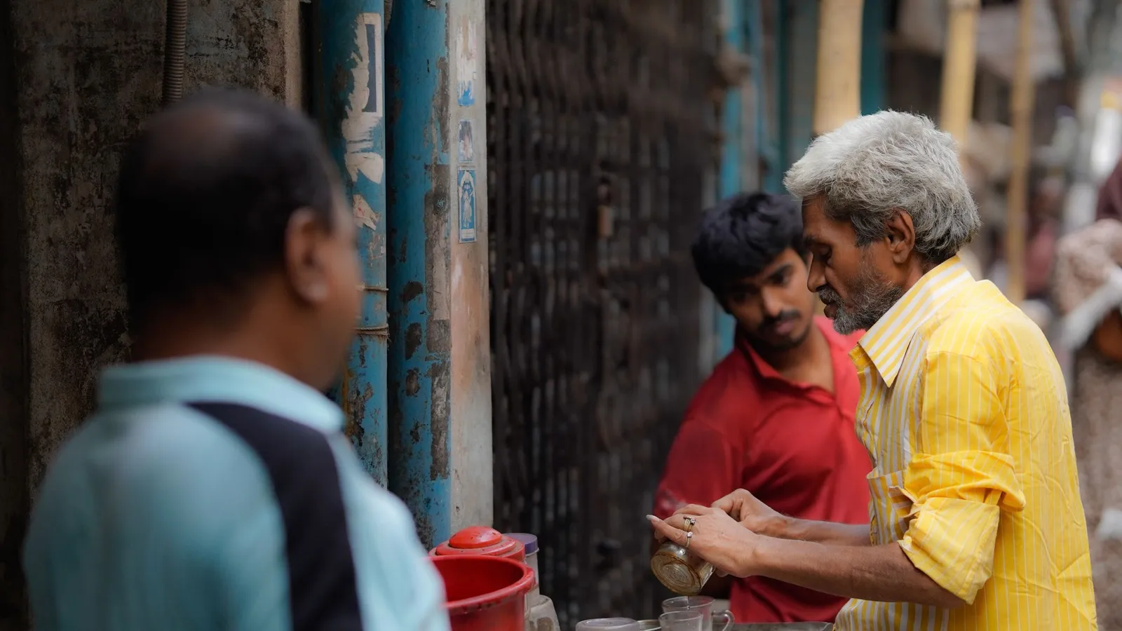 Men chat at a roadside tea stall while the vendor in a yellow shirt serves tea.