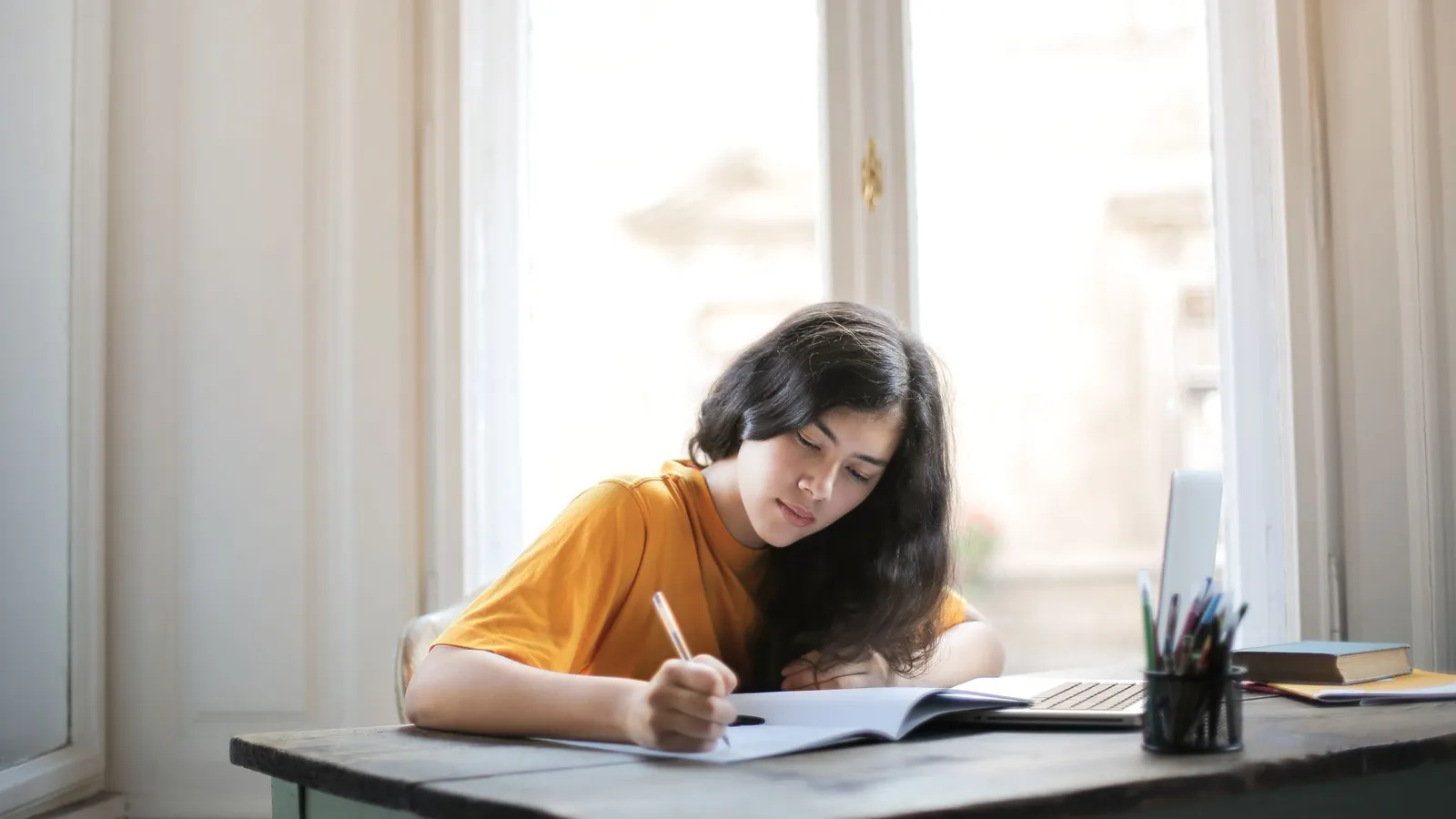 A young woman writes in a notebook at a desk beside a sunlit window.