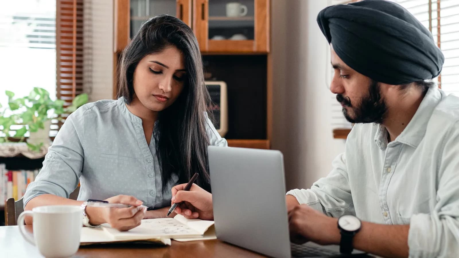 A young Indian woman and a Sikh man study papers together beside a laptop at home.