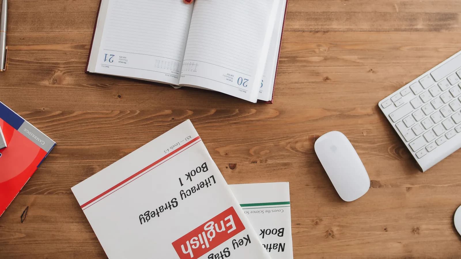 An open planner, English workbooks, a keyboard and a mouse on a wooden desk, seen from above.