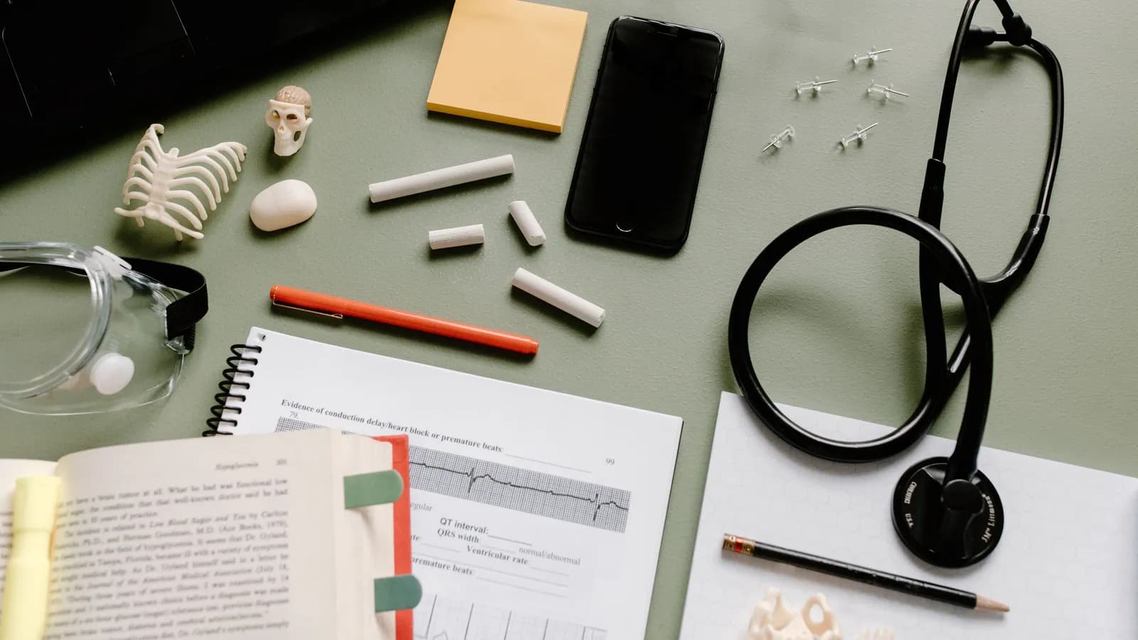 A stethoscope, study notes, pens and a phone laid out on a desk for medical study.