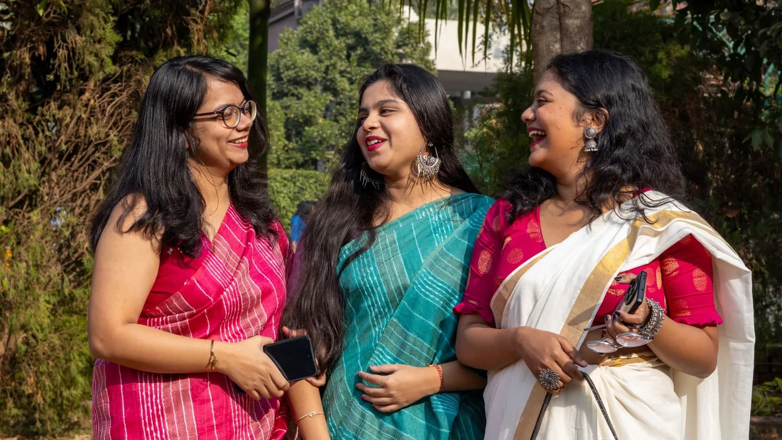 Three Indian women in saris, one in a cream-and-gold kasavu, laugh together as they talk outdoors.