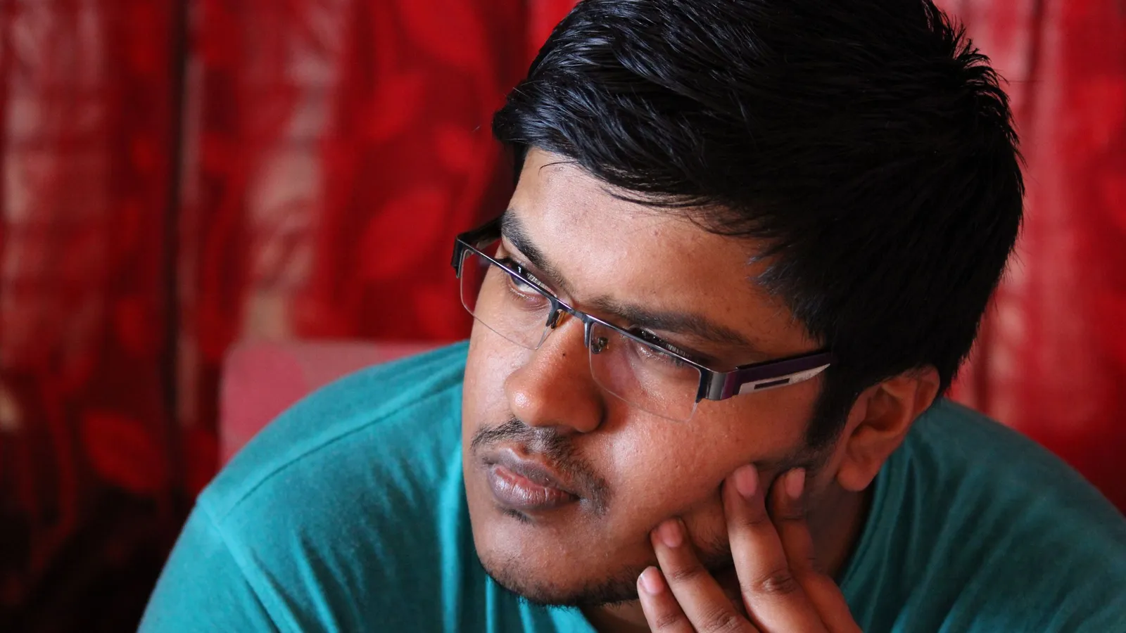 A young Indian man in glasses rests his chin on his hand, lost in thought.