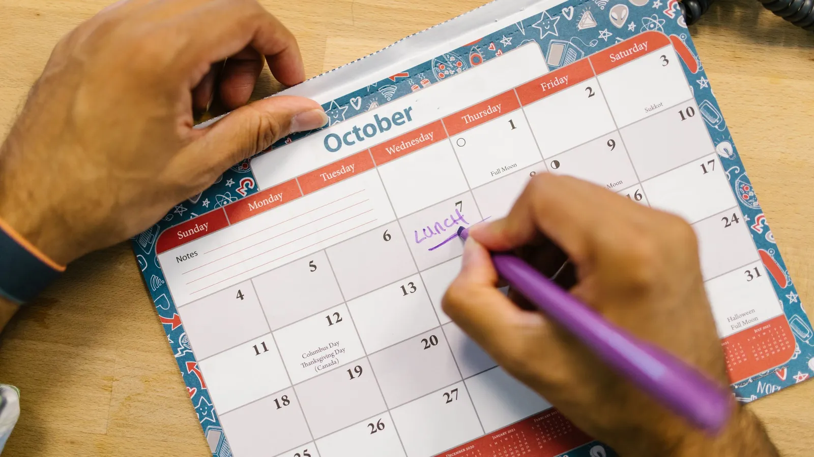 A hand writes a note on a monthly wall calendar with a purple pen.
