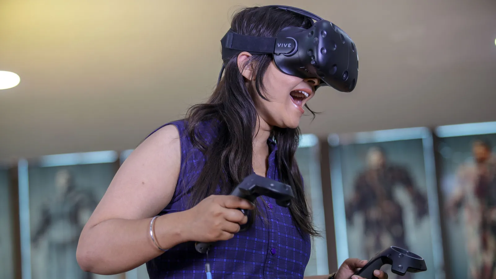 A South Asian woman wearing a VR headset holds a controller in each hand, absorbed in a virtual scene.