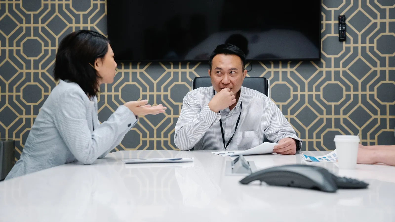 A woman explains something across a meeting table while a man listens, hand over his mouth, looking unsure.